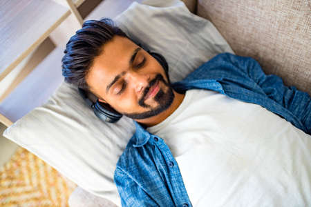 asian indian man living room lying on sofa closing his eyes and listening to music with a slight...