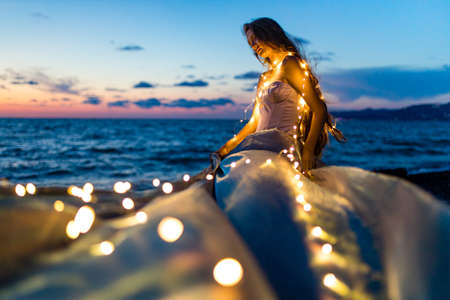 long haired woman in corset with shells on the chest looking languid in twilight near sea beach