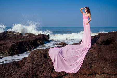 Beautiful brunette woman in pink dress with long train sitting on the rock on a beach enjoying solitude and freedomの写真素材