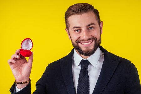 Portrait of a handsome happy man dressed in tuxedo holding open box with a ring over yellow backgroundの写真素材