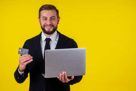 Attractive excited young brunette man isolated over yellow background holding laptop computer and plastic credit cardの写真素材