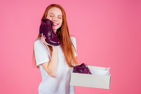 redhaired ginger woman in wtite cotton t-shirt holding burgundy lace-up boots in pink studioの写真素材