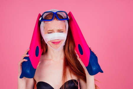 redhaired ginger woman with a bandaged face holding flippers and looking sad in studio pink backgroundの写真素材