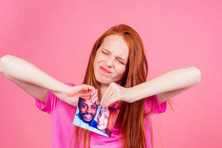 redhaired ginger woman in pink studio background tearing photo card of smiling international couple the end of the relationshipの写真素材