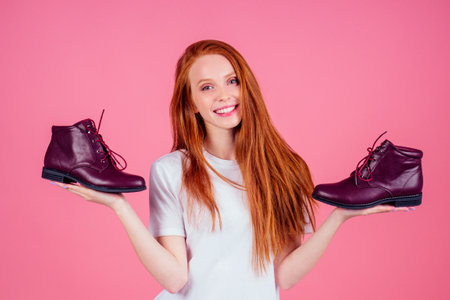 redhaired ginger woman in wtite cotton t-shirt holding burgundy lace-up boots in pink studioの写真素材