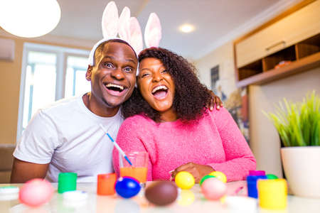 Young african american man and woman Authentic people wearing cute easter bunny ears and painting eggs in the houseの写真素材