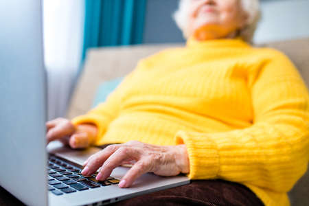 Happy mature blonde lady sitting on couch with laptop in living roomの写真素材