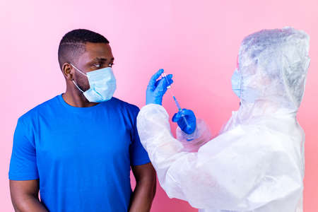 African american man scientist in PPE suite uniform showing medicine liquid vaccine vial bottle studio pink wallの写真素材