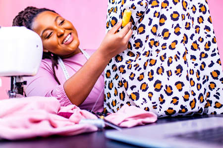 african tailor with afro dreadlocks pigtails sews clothes on sewing machine at tailor office pink...