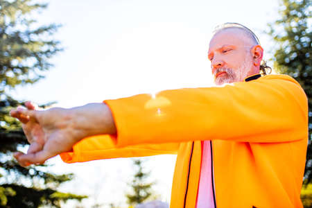 Senior man in orange sports wear doing stretching warming up outdoors sunny day in forest aloneの写真素材
