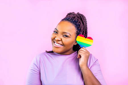 Portrait of an attractive hispanic woman holding a LGBT rainbow heart in pink studioの写真素材