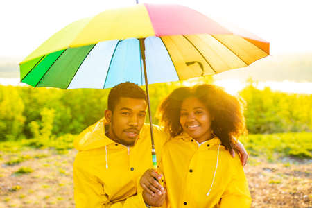 african american wife and husband in yellow raincoat with a hood feeling happy at sunset outdoors summer under rainbow umbrellaの写真素材
