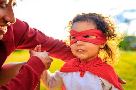 african american little girl in red costume and eyes mask playing a hero in summer park with dadの写真素材