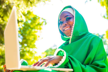 hispanic woman in green muslim hijab with bright make up and nose piercing working by laptop sit on grass outdoors summer parkの写真素材