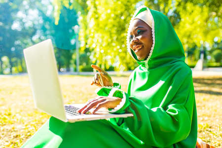 hispanic woman in green muslim hijab with bright make up and nose piercing working by laptop sit on grass outdoors summer parkの写真素材