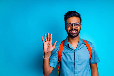 indian student man feeling happy looking at camera wear eye glasses and orange backpack and saying hello with hand in blue studioの写真素材