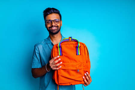 handsome young bearbed indian man with eye glasses in blue cotton t-shirt with orange rainbow backpack in studio backgroundの写真素材