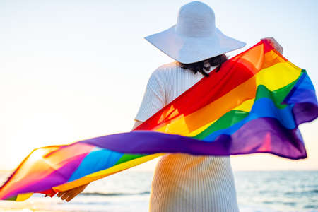 woman with straw white hat holding a gay pride rainbow flag blowing in the wind on beach with golden sunset .の写真素材