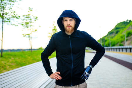 man with arm prosthesis in sports clothing ready for morning workout outdoors. Disabled sport...