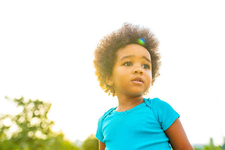 cute little african american girl in cotton blue t-shirt in spring park at sunset curly hair