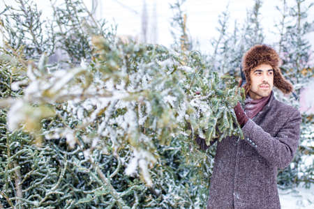 indian man in stylish wool coat buys a Christmas tree outdoors snow background