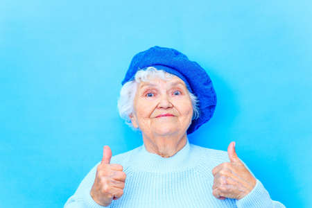 beautiful retired woman in blue beret and warm pullover looking at camera and smile in blue wall background in studioの写真素材