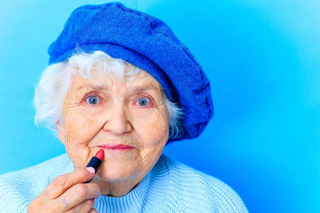 beautiful mature 80 years old woman in blue beret and woolen sweater apply lipstick in blue studio...
