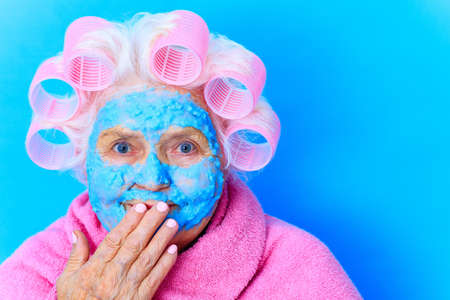 woman with blue cream moistening mask on face , puts curlers on white hair and wear pink bath robe in studio backgroundの写真素材