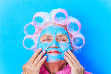 woman with blue cream moistening mask on face , puts curlers on white hair and wear pink bath robe in studio backgroundの写真素材