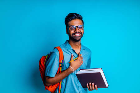handsome young bearbed indian man with eye glasses in blue cotton t-shirt with orange rainbow backpack in studio backgroundの写真素材