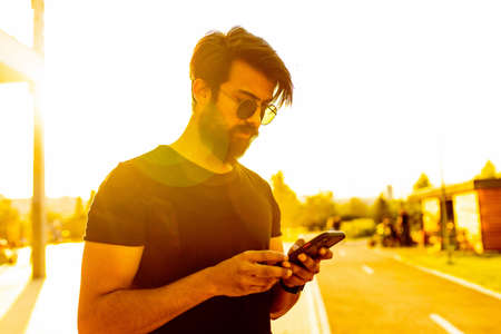 happy bearded italian man in black t-shirt useing phone in park at sunset gold lightの写真素材