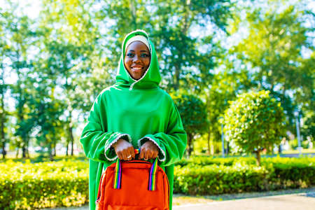 african woman in green hijab with bright makeup holding coffee to go with rainbow backpack in summer parkの写真素材