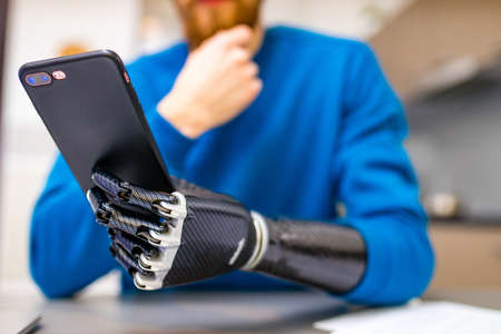 young man with bionic hand hold smartphone while sitting indoors home kitchen backgroundの写真素材