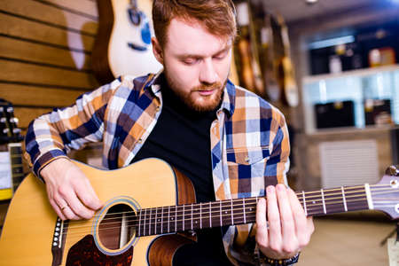 redhaired bearded handsome man in brown plaid casual shirt choosing a guitar in a music store