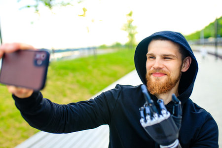 young bearded man with metal arm prosthetic outdoor taking selfie photos on smartphone cameraの写真素材