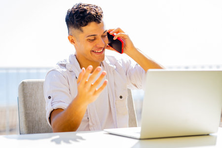 man in white t-shirt talking by phone while remote working at sunny beach in cafeの写真素材