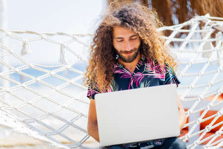 long curly haired man sitting on the hammock near the beach and working laptop vacationの写真素材