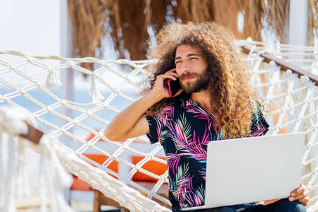 long curly haired man sitting on the hammock near the beach and working laptop vacationの写真素材