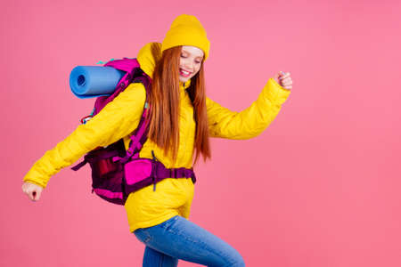 Female hiker with a backpack looking at the camera and smiling isolated on pink studio background.she wearing yellow windbreaker waterproof jacket and knitted hatの写真素材