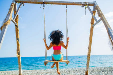 spanish mexican woman with long curly hair wear pink and blue bikini ans sinning on swing at sunny beachの写真素材