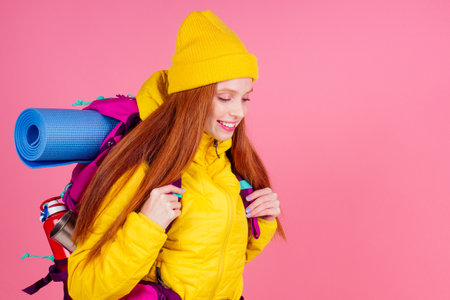 Female hiker with a backpack looking at the camera and smiling isolated on pink studio background.she wearing yellow windbreaker waterproof jacket and knitted hatの写真素材