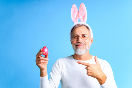 Cute mature man with bunny ears holding Easter egg on blue color backgroundの写真素材