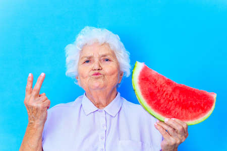 mature woman with snow white grey white hair in blue shirt holding watermelon in studio backgroundの写真素材
