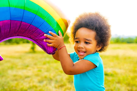 positive multicultural race little girl with afro curly hair holding rainbow balloon in summer parkの写真素材