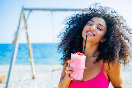 latin brazilian woman with curly long hairstyle and pink bikini drinking strawberry smoothie in beach on the swingの写真素材