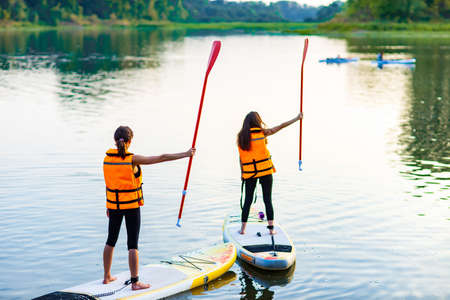 two women in life jacket at sub board at river ar evening , forest trees background