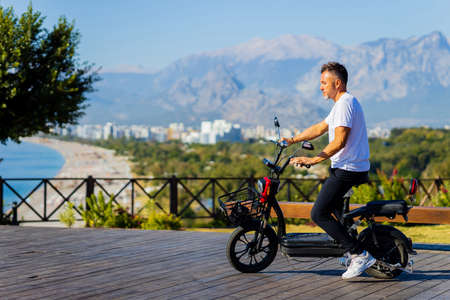 handsome man in white t-shirt sitting on the bench next to eco scooter with beautiful view on hillsの写真素材