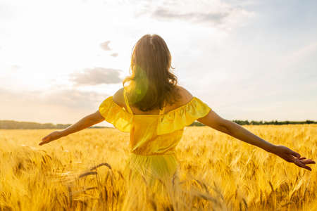Young woman in yellow dress standing on a wheat field with sunrise on the backgroundの写真素材