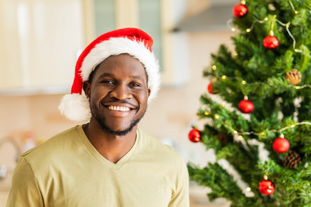 afro american man with snow white smile in Santa hat looking at camera near christmas tree at homeの写真素材