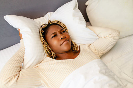 african american woman trying to sleep but the noise bothers her she is covering the ears with a pillowの写真素材
